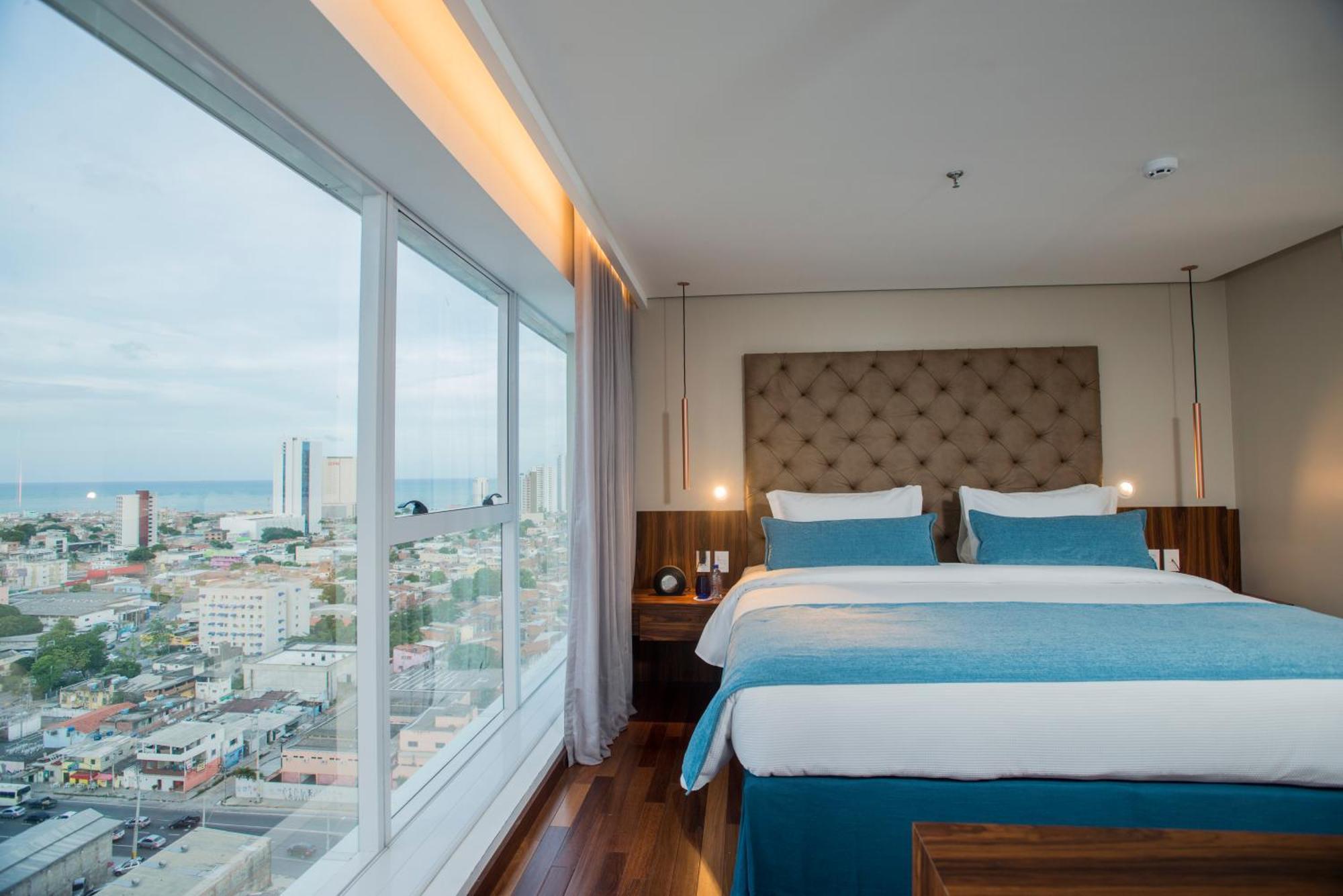 Hotel Luzeiros Recife Zewnętrze zdjęcie The photo shows a modern hotel room featuring a large window with a city view. Inside, there are two neatly made beds with light blue and white bedding, flanked by wooden nightstands. The room has a contemporary design, with a padded headboard and so